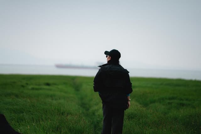 A photograph taken from behind of a person standing alone in a wide open green field under an overcast gray sky. The person wears a black cap, black jacket, and dark pants, with sunglasses visible in profile. They face toward the distant horizon where a body of water meets the sky, with a large ship faintly visible on the water. A faint path cuts through the grass in front of them.
