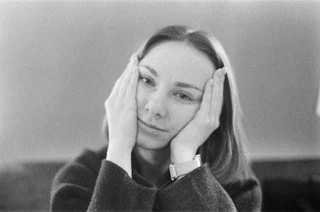Alt Text: A black and white film-style photograph of a young woman with straight shoulder-length hair sitting with both hands raised to the sides of her face, fingers touching her temples and cheeks. Her expression is contemplative and weary. She wears a textured dark sweater and a watch on her left wrist. The background is a softly blurred neutral gray.