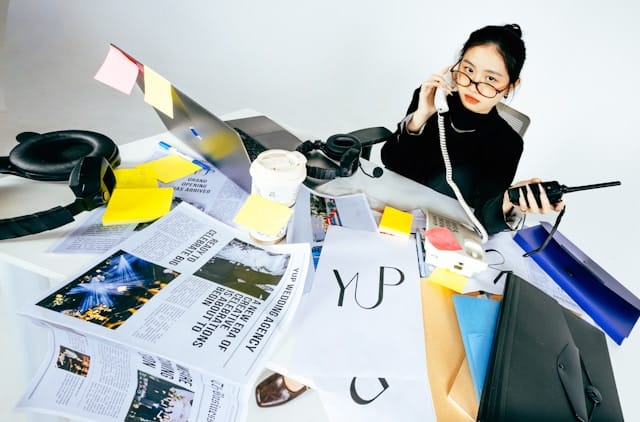 An overhead photograph of a young East Asian woman with her hair up, wearing a black turtleneck and glasses, seated at a desk completely covered in overlapping newspapers, yellow sticky notes, folders, a coffee cup, headphones, and electronic devices including a corded phone and a walkie-talkie. She holds the corded phone to her ear with one hand and a device in the other. Her expression is calm but surrounded by visible chaos.