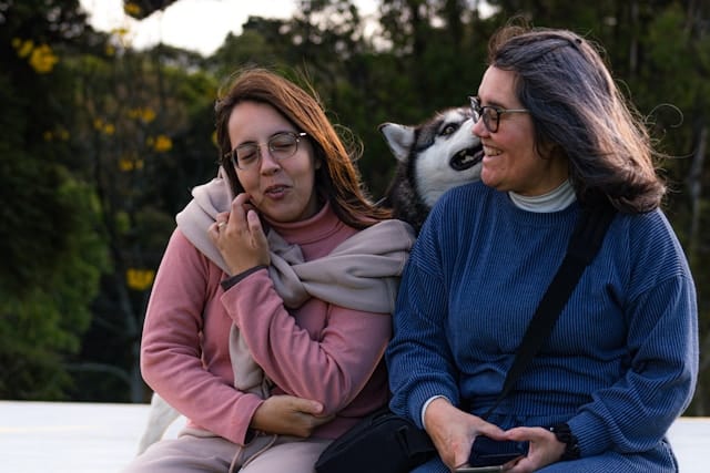 Two women having a warm conversation outdoors with a husky dog, surrounded by trees in a natural park setting