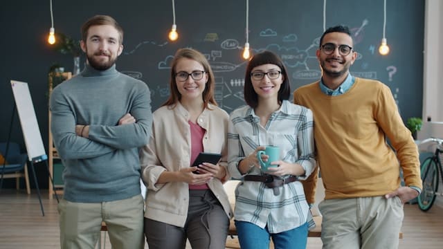 Four diverse young professionals standing together smiling in modern office with chalkboard and pendant lights in background
