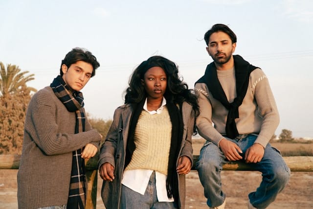 Three diverse young adults sitting outdoors, with woman in yellow sweater seated between two men wearing neutral colored clothing