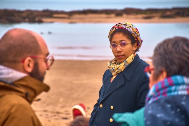 Woman wearing colorful patterned headscarf and glasses standing on beach with two people facing her in conversation