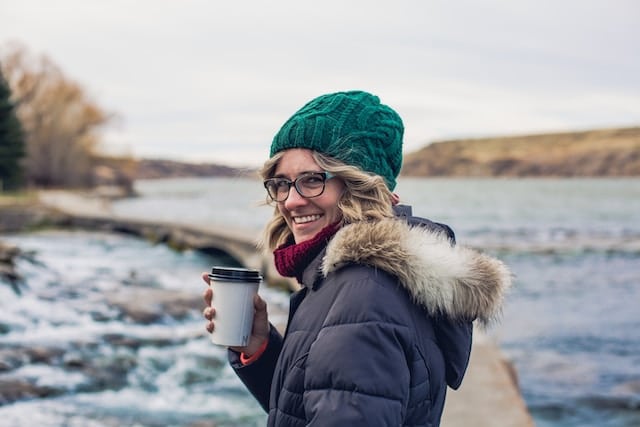 Smiling blonde woman wearing green knit hat and winter coat holding coffee cup outdoors by water