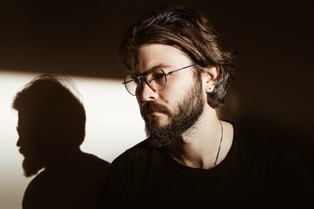 Bearded man wearing glasses in contemplative pose with dramatic shadow on wall behind him