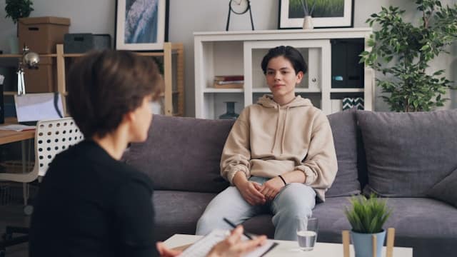 Therapist taking notes while sitting across from young client on gray couch in comfortable therapy office
