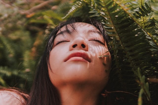 Woman with eyes closed in peaceful expression, her face dappled with light filtering through surrounding fern fronds
