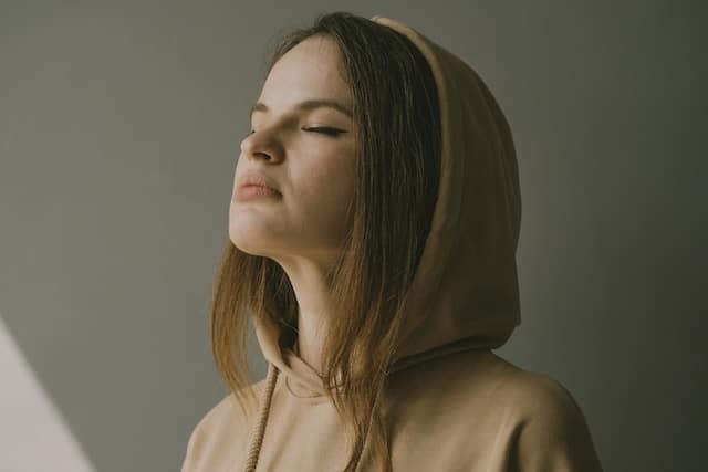 Young woman wearing a tan hoodie with eyes closed in a calm meditative expression, photographed in soft natural light against a neutral background