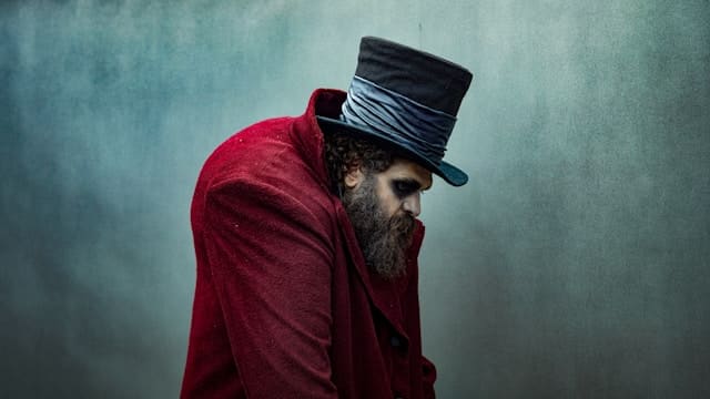 Person wearing red jacket and black top hat with head bowed and shoulders curved forward against teal background