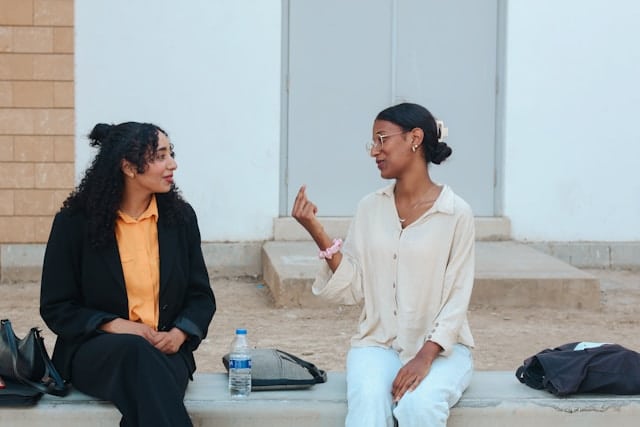 Two women sitting on outdoor steps engaged in friendly conversation, one with curly hair in a black jacket, the other wearing glasses and a cream colored shirt