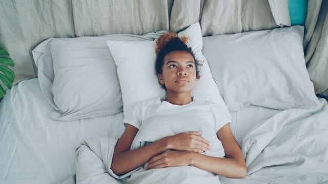 Woman with curly hair lying in white bed awake staring at ceiling with hands folded on chest