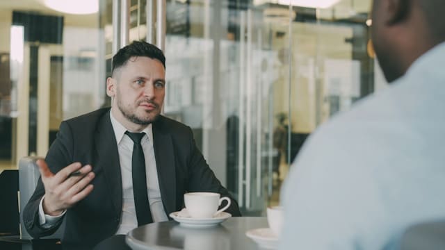 Two men seated at a table having an engaged conversation, one gesturing while speaking, with coffee cups between them