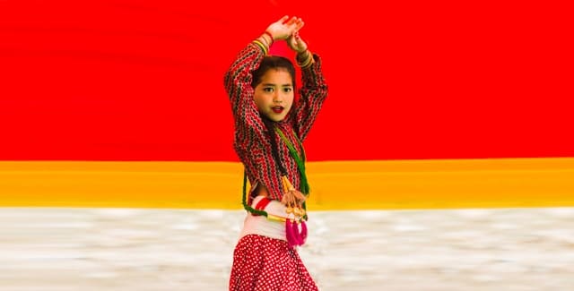 Young girl in traditional red and yellow cultural dress performing dance pose against vibrant background