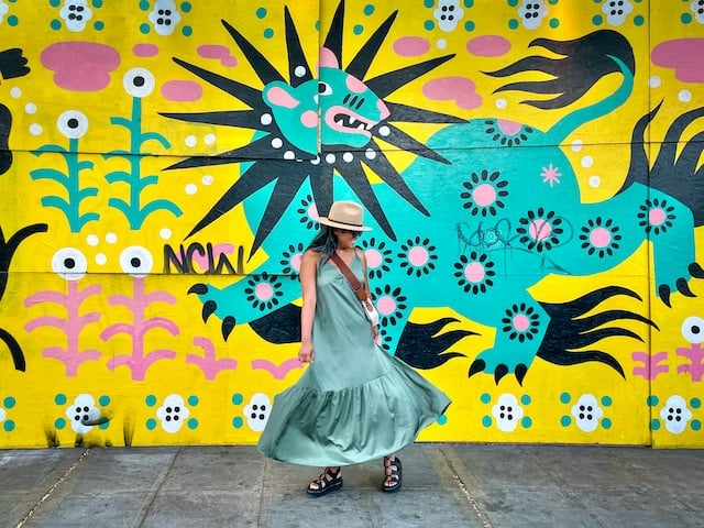 Woman in flowing turquoise dress walking past vibrant yellow and blue street mural