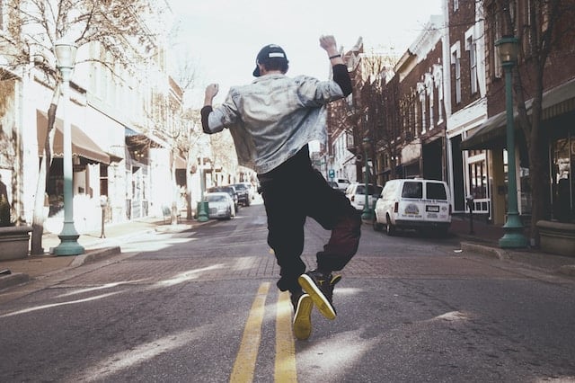 Person jumping joyfully on small town street, wearing denim jacket and dark pants