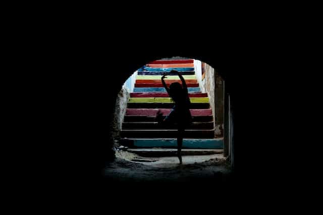 Silhouette of dancer in ballet pose inside dark archway with colorful rainbow painted stairs beyond