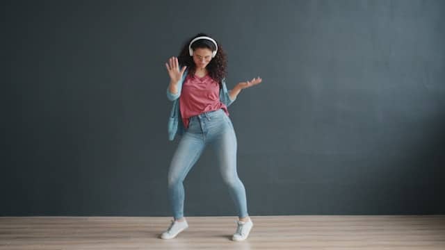 Young woman with headphones dancing against gray wall, wearing casual denim and pink top