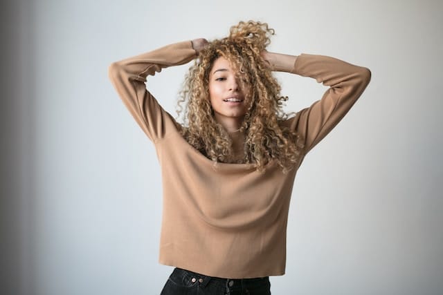 Young woman with curly blonde hair wearing a tan sweater, both hands in her hair with an expression of stress or overwhelm.
