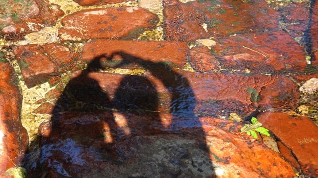 Shadow of two people making a heart shape with their hands together, cast on wet reddish stones with shallow water
