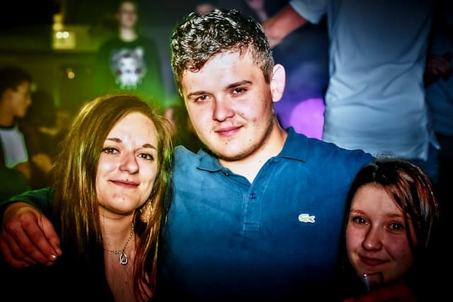Three young adults standing close together with arms around each other at a social gathering, smiling at the camera