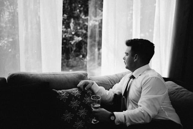 Black and white photograph of a man in a white dress shirt sitting on a couch near sheer curtains, looking pensively out a window while holding a glass.