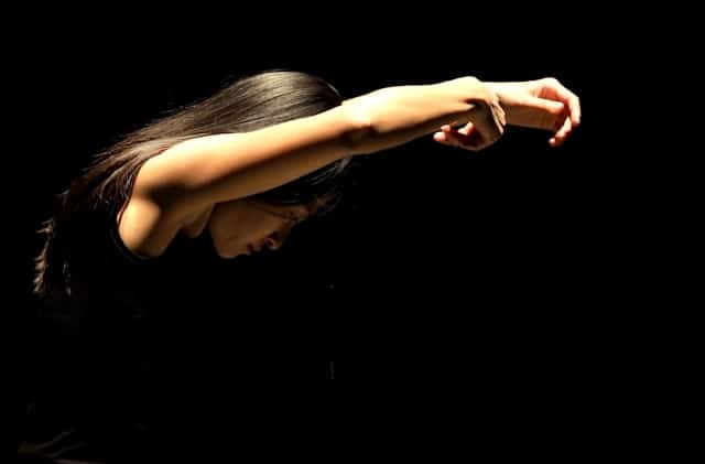 Woman with long dark hair extending her arms forward against a black background in an expressive pushing gesture, face partially obscured