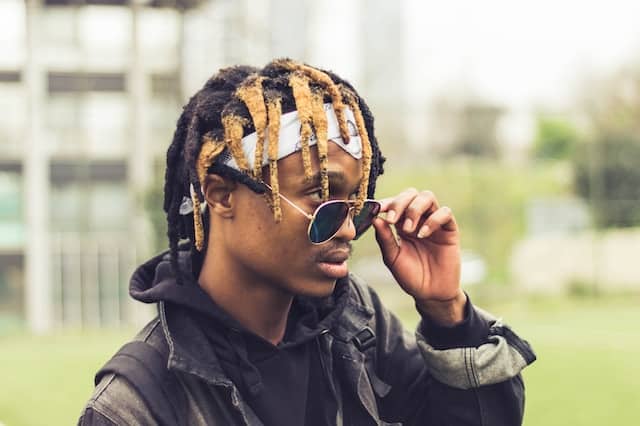 Young Black man with golden dreadlocks lowering his sunglasses and looking thoughtfully to the side in an urban outdoor setting.