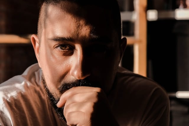 Close up portrait of a bearded man with his hand near his chin, half of his face illuminated while the other half remains in shadow.