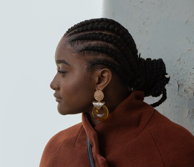 Black woman with braided hair styled in an updo, wearing gold earrings and a rust-colored turtleneck, shown in contemplative side profile.