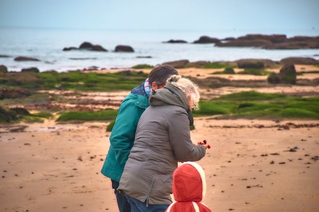 Adult and two children in winter jackets walking together on a beach with rocky shoreline and ocean in the background, viewed from behind