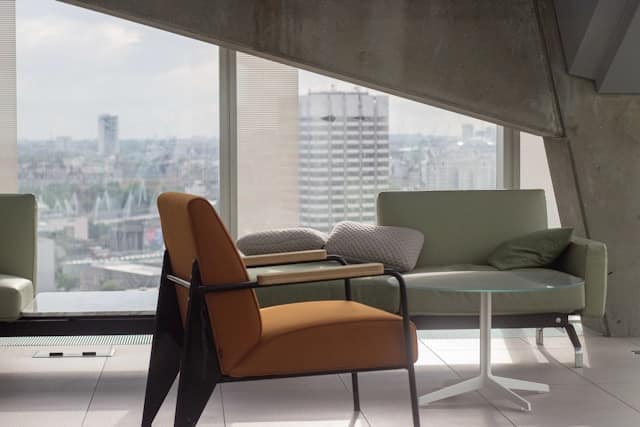 Modern seating area with an orange and brown mid century chair and green sofa positioned near large windows overlooking an urban cityscape.