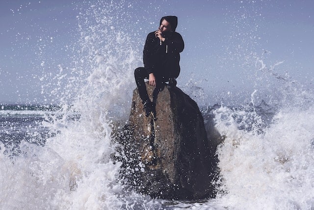 Person in dark clothing sitting calmly on a large rock while ocean waves crash dramatically around them, symbolizing finding stability amid emotional turbulence.