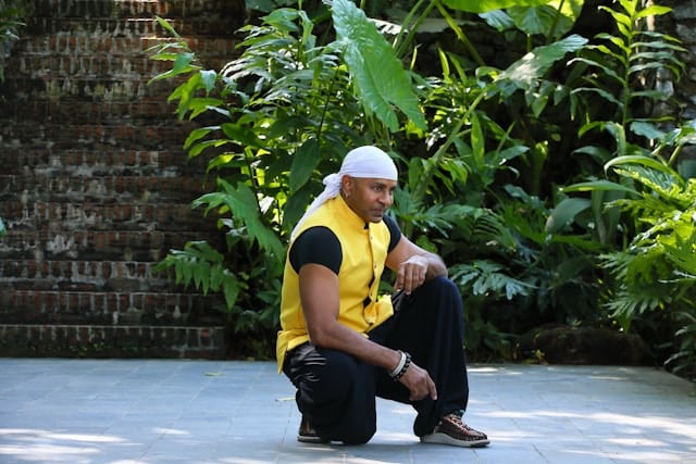 Man in yellow vest crouching in a grounded position on stone pavement surrounded by lush tropical plants against a brick wall