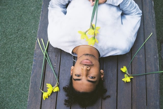 Person lying peacefully on a wooden deck surrounded by yellow daffodils, wearing a light colored shirt with eyes gently closed.