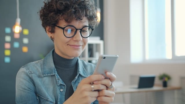 A woman with short curly hair and glasses smiling while using her smartphone in a bright office space.