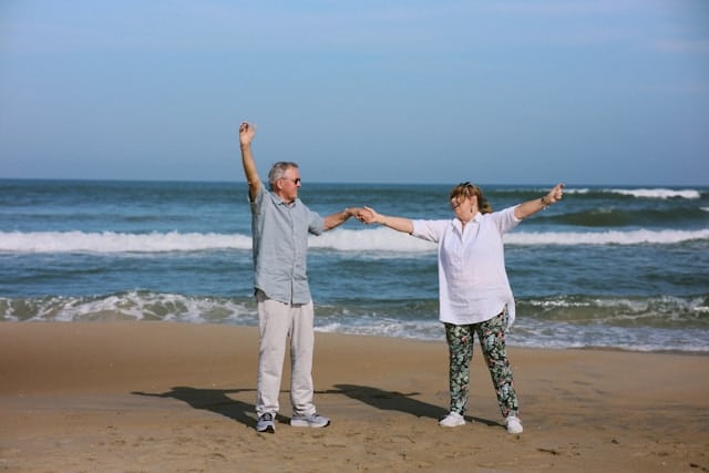 Older couple holding hands and raising their arms while moving together on a sandy beach with ocean waves in the background