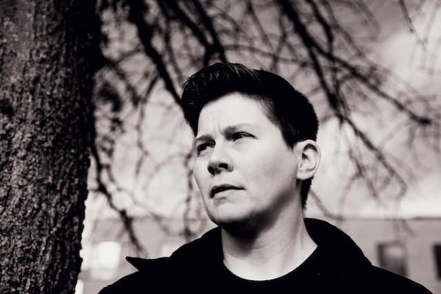 Person with short dark hair standing outdoors near bare tree branches, looking upward with a thoughtful and contemplative expression.