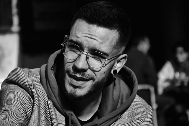 Black and white portrait of a young man wearing glasses and a hoodie under a blazer, looking slightly to the side with a contemplative expression