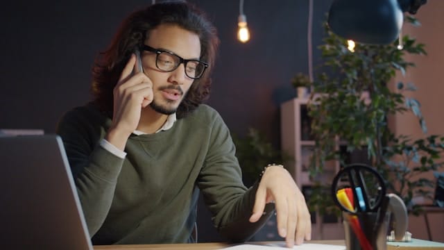 A man with long hair and glasses speaking on the phone at a desk, appearing focused and stressed.