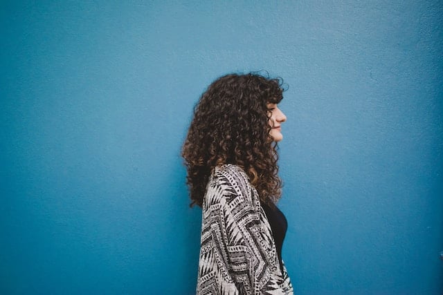 oman with curly dark hair in profile view wearing a patterned top, standing peacefully against a bright blue background.