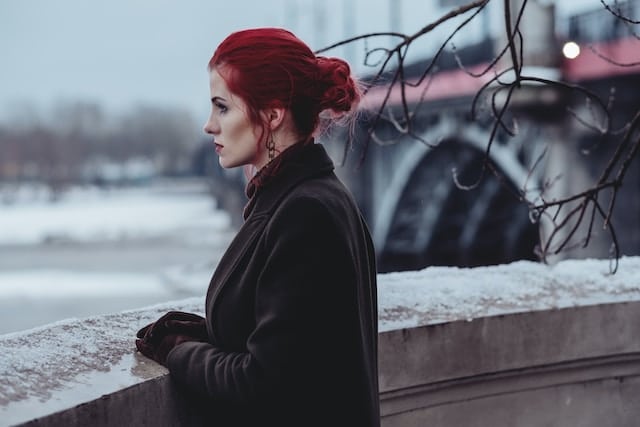 Woman with vibrant red hair in an updo wearing a dark coat, gazing pensively across a winter river scene with a bridge in the background.
