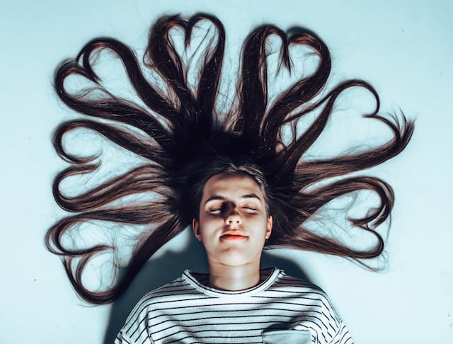 Woman lying on her back on a light blue surface with long dark hair artistically arranged in heart shapes around her head, eyes closed in peaceful relaxation.