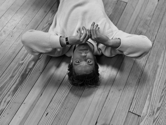 Black and white photograph of person lying on wooden floor with hands resting gently on their forehead, looking upward with a calm expression