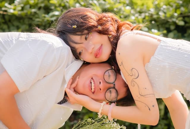 Two young women in white tops sharing a joyful moment outdoors, one playfully leaning her head on the other who is laughing, surrounded by green foliage