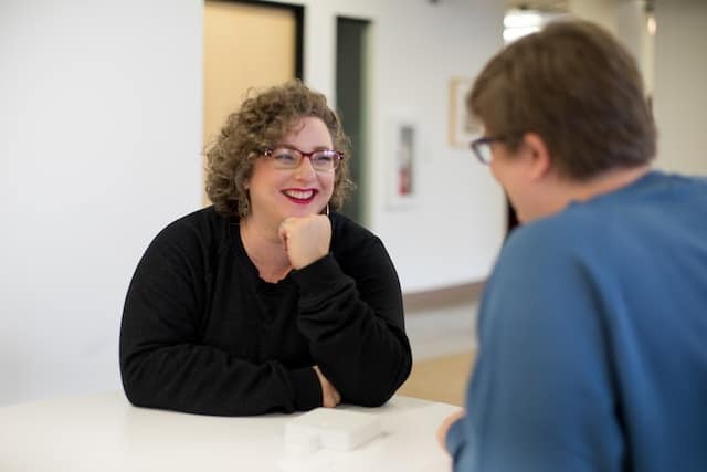 Two people sitting across from each other at a table during a therapy session, therapist wearing black and client wearing blue, engaged in conversation.