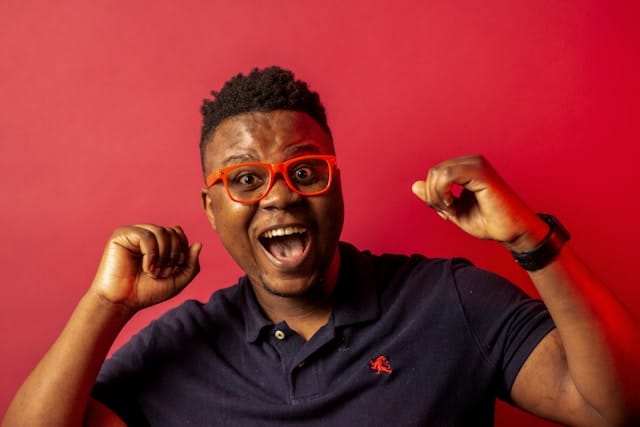 A person in a blue pop shirt wearing red glasses against a red background with an exaggerated joyful expression