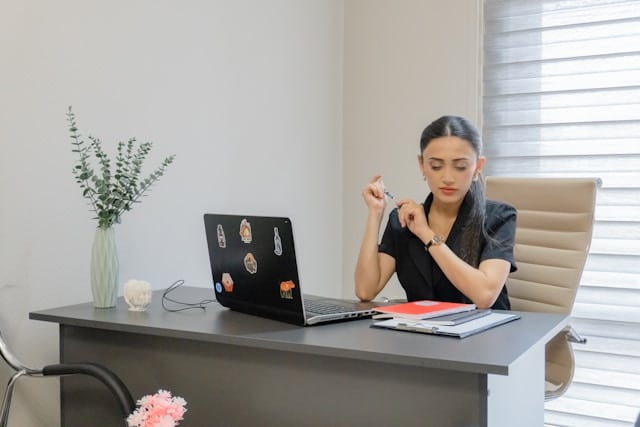 A woman sitting at a desk wearing a black shirt, with a laptop open in front of her, looking worried.