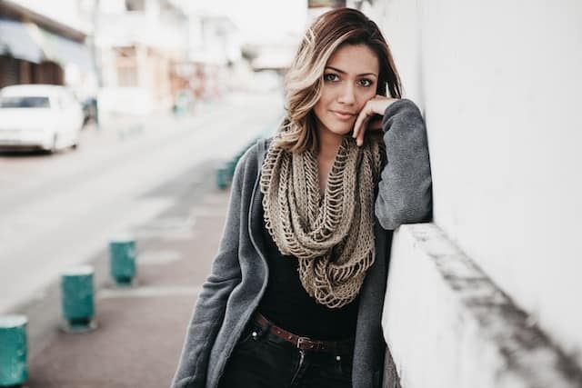 Woman with wavy brown hair wearing a gray coat and knit scarf leaning gently against a white wall on a quiet city street.