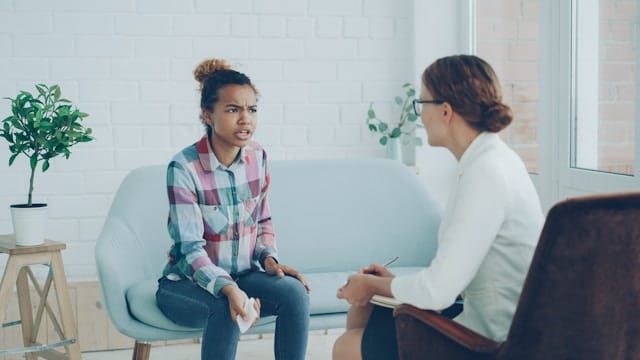 Young woman with curly hair in a bun wearing a plaid shirt sitting on a light blue couch, talking to a female therapist in white who is holding a notepad.