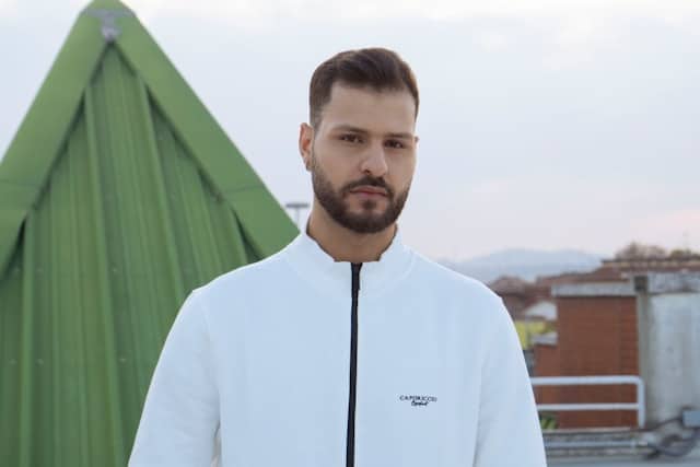 Portrait of a young man with short brown hair and a beard, wearing a white zip up athletic jacket with a small logo. He stands outdoors with a green industrial structure and city rooftops visible in the blurred background. His expression is neutral and direct, conveying approachability and readiness. The overcast sky creates soft, even lighting across his face.